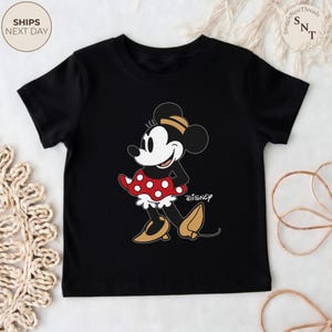 May include: Black t-shirt featuring a cartoon Minnie Mouse design. Minnie Mouse is depicted in her classic outfit: a red polka-dot skirt, yellow shoes, and a matching bow. The Disney logo is visible. The shirt is a children's apparel item.