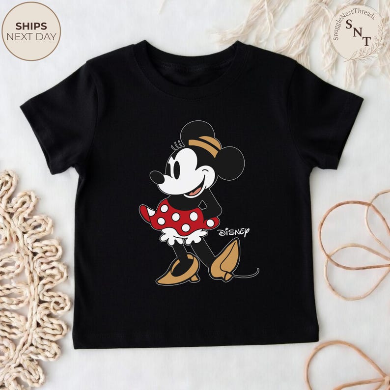 May include: Black t-shirt featuring a cartoon Minnie Mouse design. Minnie Mouse is depicted in her classic outfit: a red polka-dot skirt, yellow shoes, and a matching bow. The Disney logo is visible. The shirt is a children's apparel item.