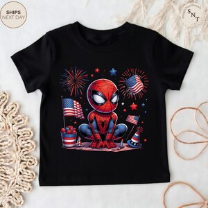 May include: A black short-sleeve t-shirt featuring a graphic of a cartoon Spider-Man character crouching. The design includes American flags, red and blue fireworks, a bucket of berries, and a party hat, all in a patriotic red, white, and blue theme.