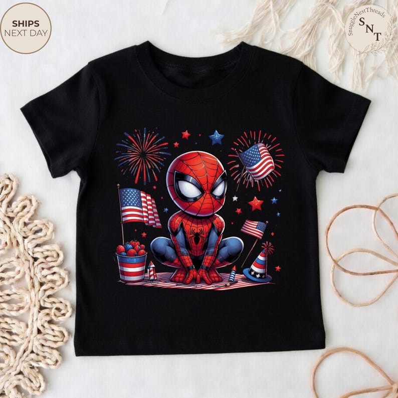 May include: A black short-sleeve t-shirt featuring a graphic of a cartoon Spider-Man character crouching. The design includes American flags, red and blue fireworks, a bucket of berries, and a party hat, all in a patriotic red, white, and blue theme.