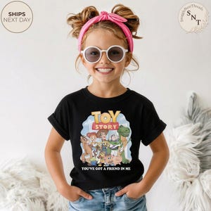 K&ouml;nnte beinhalten: Ein schwarzes T-Shirt mit Toy Story-Figuren, dem Text "TOY STORY" und "YOU'VE GOT A FRIEND IN ME". Das T-Shirt wird von einem Kind mit Sonnenbrille und einem rosa Stirnband getragen.