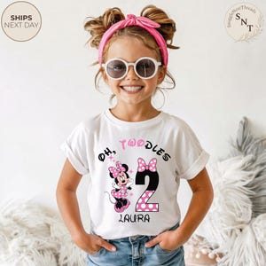 Puede incluir: Una camiseta blanca con un dise&ntilde;o de Minnie Mouse, el n&uacute;mero 2 y el texto "Oh, Twoodles" en negro y rosa. El nombre "Laura" est&aacute; impreso debajo del n&uacute;mero 2. La ni&ntilde;a lleva gafas de sol y una diadema rosa.