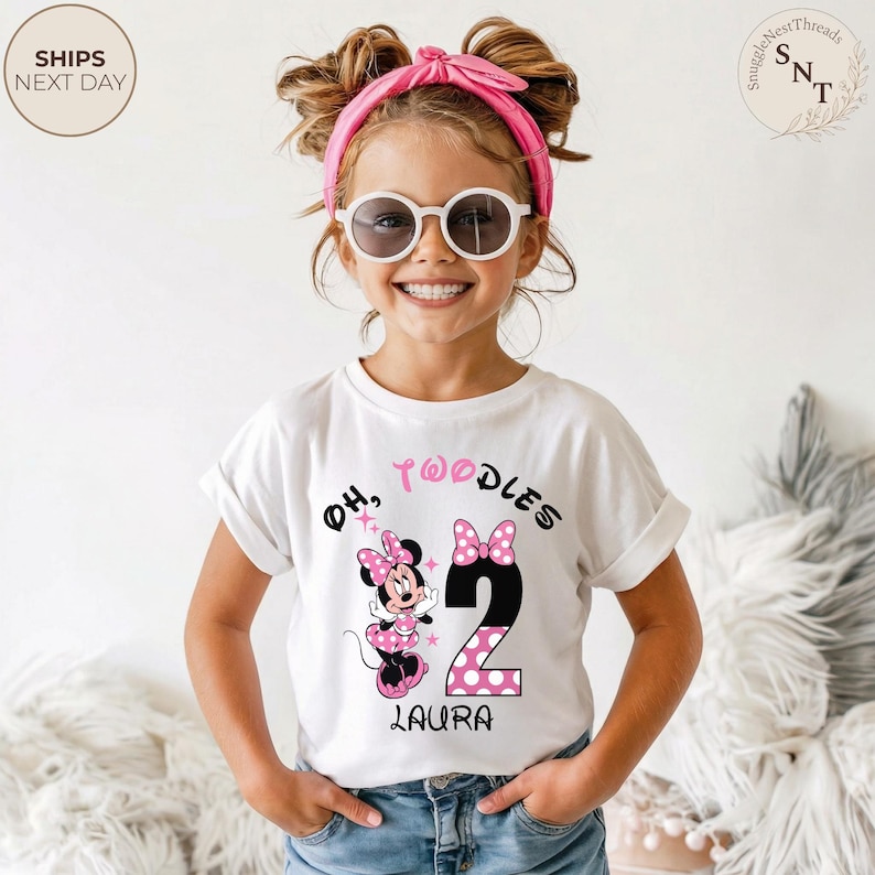 Puede incluir: Una camiseta blanca con un dise&ntilde;o de Minnie Mouse, el n&uacute;mero 2 y el texto "Oh, Twoodles" en negro y rosa. El nombre "Laura" est&aacute; impreso debajo del n&uacute;mero 2. La ni&ntilde;a lleva gafas de sol y una diadema rosa.