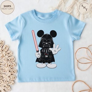 May include: Light blue toddler t-shirt featuring a cartoon mashup of Darth Vader and Mickey Mouse. The character is black with a red lightsaber and white gloves and shoes. The shirt has the text "SHIPS NEXT DAY" in a circle.
