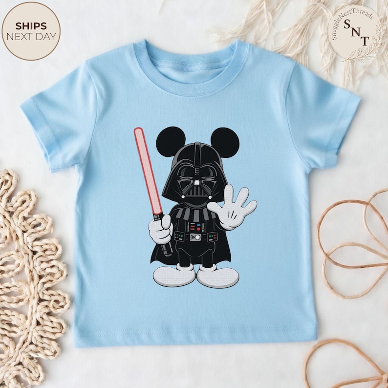 May include: Light blue toddler t-shirt featuring a cartoon mashup of Darth Vader and Mickey Mouse. The character is black with a red lightsaber and white gloves and shoes. The shirt has the text "SHIPS NEXT DAY" in a circle.