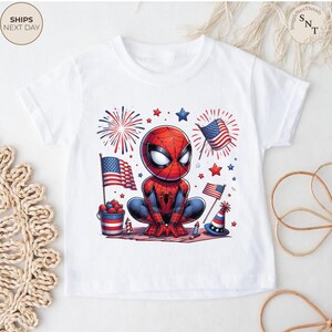 May include: A white short-sleeve t-shirt featuring a graphic of a cartoon Spider-Man character crouching. The design includes American flags, red and blue fireworks, a bucket of berries, a party hat, and stars in a patriotic red, white, and blue color scheme.