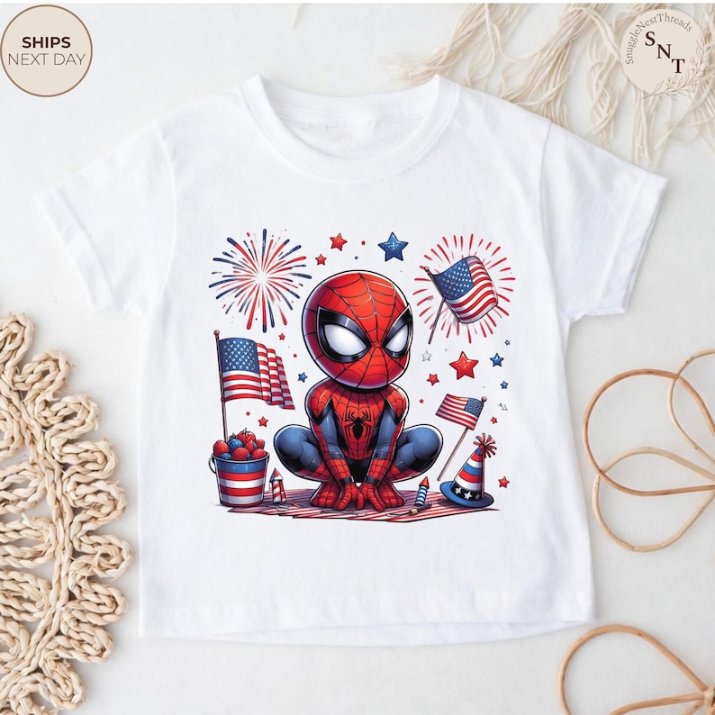 May include: A white short-sleeve t-shirt featuring a graphic of a cartoon Spider-Man character crouching. The design includes American flags, red and blue fireworks, a bucket of berries, a party hat, and stars in a patriotic red, white, and blue color scheme.