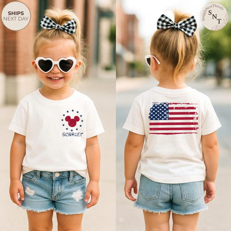 May include: A young child wearing a white t-shirt with a red, white, and blue design. The front of the shirt features a Mickey Mouse head and the name "Scarlet". The back of the shirt displays a distressed American flag. The child is also wearing denim shorts and heart-shaped sunglasses.