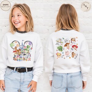 Puede incluir: Una sudadera blanca de manga larga con un gr&aacute;fico frontal de personajes de Toy Story dispuestos en forma de orejas de Mickey Mouse con un castillo, y un gr&aacute;fico trasero que muestra caras de personajes como Buzz, Woody y Jessie con nombres.