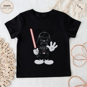 May include: Black t-shirt featuring a cartoon character mashup of Darth Vader and Mickey Mouse. The character holds a red lightsaber and waves with a white gloved hand. The shirt has a round neck and short sleeves. The text "SHIPS NEXT DAY" is in the upper left corner.