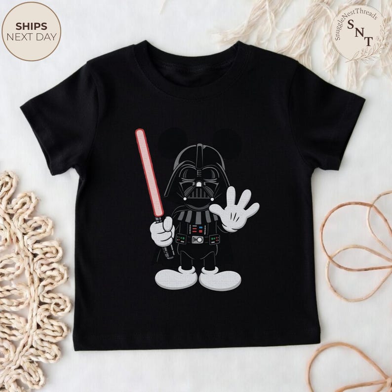 May include: Black t-shirt featuring a cartoon character mashup of Darth Vader and Mickey Mouse. The character holds a red lightsaber and waves with a white gloved hand. The shirt has a round neck and short sleeves. The text "SHIPS NEXT DAY" is in the upper left corner.
