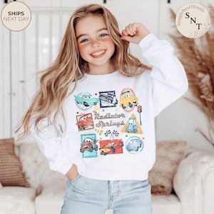 May include: White sweatshirt featuring a colorful graphic of cartoon cars and the text "Radiator Springs." The design includes various car illustrations within postage stamp-like frames. The sweatshirt has long sleeves and a crew neck.