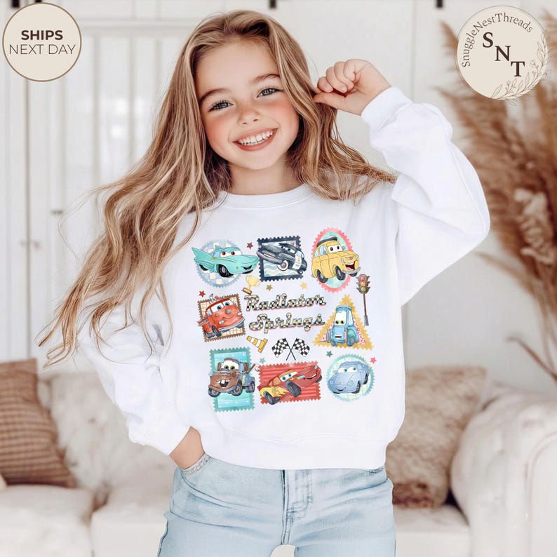 May include: White sweatshirt featuring a colorful graphic of cartoon cars and the text "Radiator Springs." The design includes various car illustrations within postage stamp-like frames. The sweatshirt has long sleeves and a crew neck.