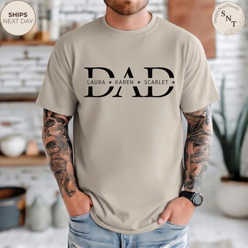 Puede incluir: Camiseta beige con la palabra "DAD" en letras negras grandes. Debajo de la palabra "DAD" est&aacute;n los nombres "LAURA", "KAREN" y "SCARLET" con estrellas. La camiseta tiene mangas cortas y cuello redondo.