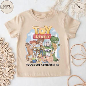 K&ouml;nnte beinhalten: Beiges kurz&auml;rmeliges T-Shirt mit dem Schriftzug "TOY STORY" in einem farbenfrohen Design, mit Zeichentrickfiguren aus dem Film. Der Satz "YOU'VE GOT A FRIEND IN ME" ist darunter gedruckt. Das T-Shirt hat einen Rundhalsausschnitt.