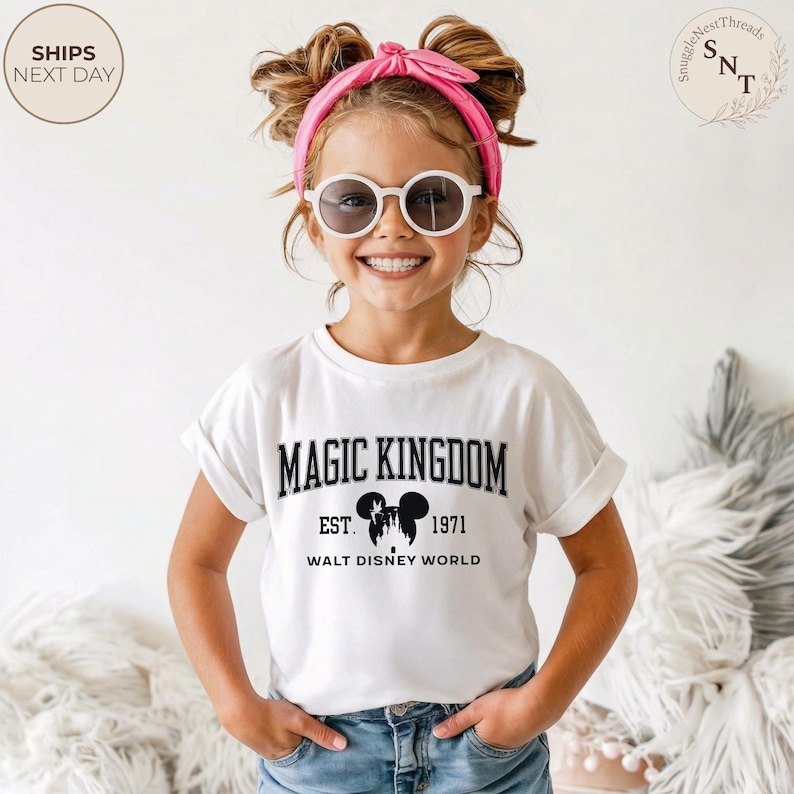 May include: A white t-shirt with the words "MAGIC KINGDOM EST. 1971 WALT DISNEY WORLD" in black. The shirt features a graphic of Mickey Mouse. The child is wearing sunglasses and a pink headband.