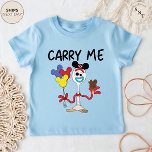 May include: A light blue toddler t-shirt with the words "CARRY ME" in black. The shirt features a cartoon character with a spoon for a body, holding balloons and a chocolate treat. The character is wearing a Mickey Mouse hat.