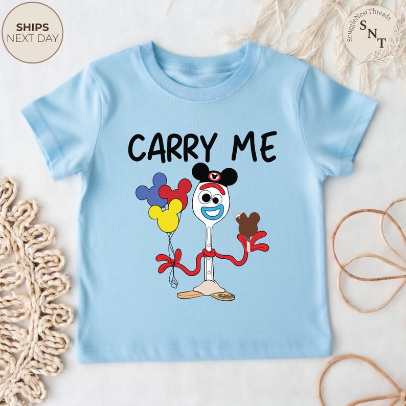 May include: A light blue toddler t-shirt with the words "CARRY ME" in black. The shirt features a cartoon character with a spoon for a body, holding balloons and a chocolate treat. The character is wearing a Mickey Mouse hat.