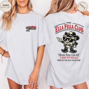 Puede incluir: Camiseta azul claro con un gr&aacute;fico de "THE ELLA FELLA CLUB" en la espalda, con un vaso de whisky de dibujos animados con sombrero y botas de vaquero. Peque&ntilde;o logo en la parte delantera. Texto: "Choosin' Texas, I can tell."