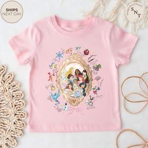 May include: A light pink t-shirt featuring a gold-framed portrait of Disney princesses. Surrounding the portrait are various character illustrations and signatures. The shirt has short sleeves and a crew neck.