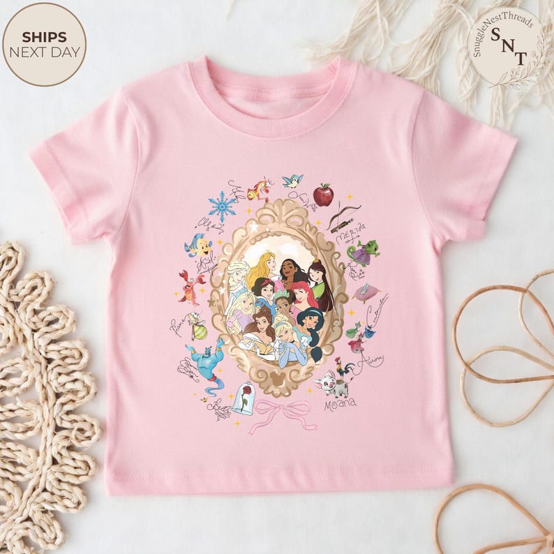 May include: A light pink t-shirt featuring a gold-framed portrait of Disney princesses. Surrounding the portrait are various character illustrations and signatures. The shirt has short sleeves and a crew neck.