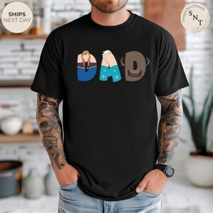 K&ouml;nnte beinhalten: Schwarzes T-Shirt mit dem Wort "DAD" im Cartoon-Stil. Das "D" ist braun mit Geweih, das "A" ist blau mit Schneeflocken und das erste "D" ist blau und lila mit einem Zopf. Das Shirt hat den Text "SnuggleNestThreads".