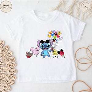 Pu&ograve; includere: T-shirt bianca con un'illustrazione di Stitch che tiene in mano palloncini, zucchero filato e gelato. Il design include elementi a tema Mickey Mouse. La maglietta ha il testo "SHIPS NEXT DAY" in alto a sinistra.