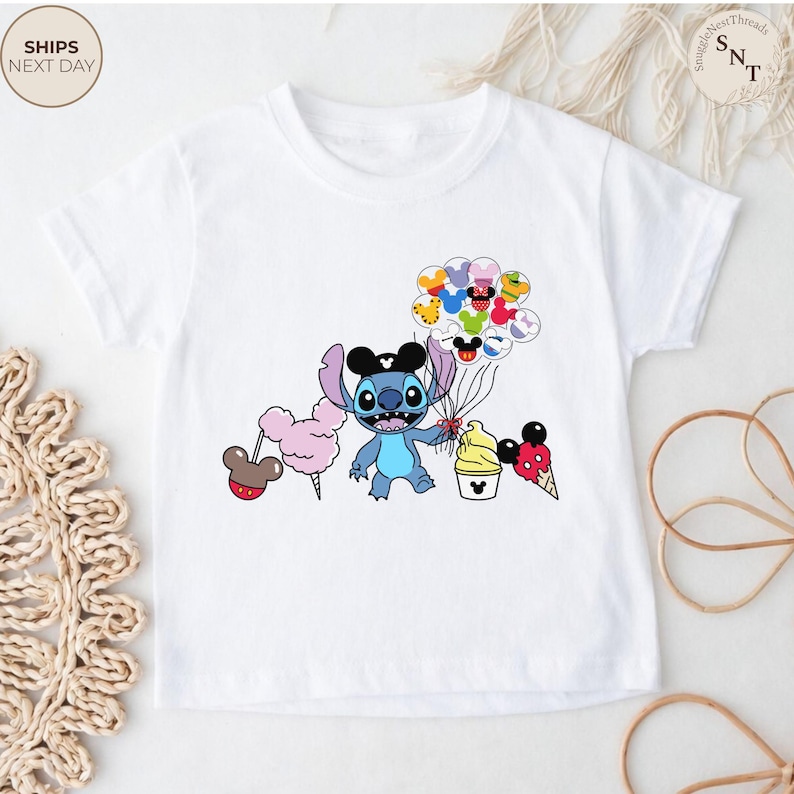 Pu&ograve; includere: T-shirt bianca con un'illustrazione di Stitch che tiene in mano palloncini, zucchero filato e gelato. Il design include elementi a tema Mickey Mouse. La maglietta ha il testo "SHIPS NEXT DAY" in alto a sinistra.