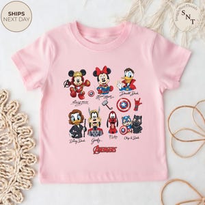 May include: A light pink t-shirt featuring a colorful graphic of various Disney characters in the Avengers theme. The characters include Mickey Mouse, Minnie Mouse, Donald Duck, Daisy Duck, Goofy, Pluto, and Chip & Dale. The word "AVENGERS" is printed in red.