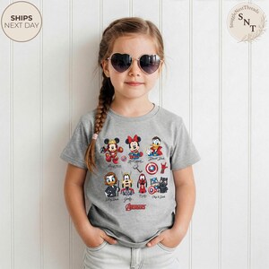 May include: A young girl wearing heart-shaped sunglasses and a gray t-shirt featuring Disney characters as Avengers. The shirt displays Mickey Mouse, Minnie Mouse, Donald Duck, and other characters with the Avengers logo.