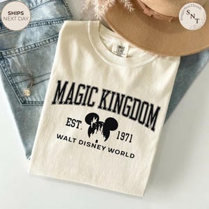 May include: Cream-colored t-shirt with the text "MAGIC KINGDOM" in black arched lettering. Below the text is "EST. 1971" and "WALT DISNEY WORLD". The shirt features a Mickey Mouse silhouette. The shirt is laid on top of blue jeans.