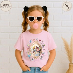 May include: A light pink t-shirt with a gold-framed illustration of Disney princesses. The shirt is worn by a child wearing heart-shaped sunglasses and a pink bubblegum bubble. The shirt has the text "SnuggleNestThreads" and "Ships Next Day".
