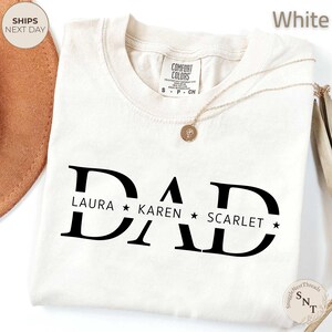 Puede incluir: Camiseta blanca con la palabra "DAD" en letras negras grandes, con los nombres "LAURA", "KAREN" y "SCARLET" debajo. Un collar dorado con un colgante est&aacute; sobre la camiseta. La camiseta tiene una etiqueta "Comfort Colors".