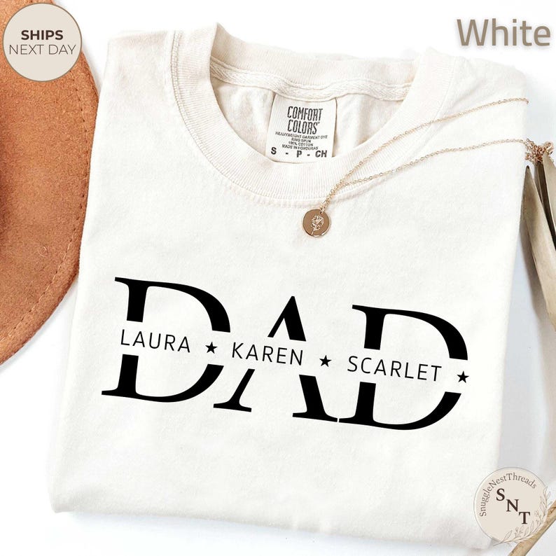 Puede incluir: Camiseta blanca con la palabra "DAD" en letras negras grandes, con los nombres "LAURA", "KAREN" y "SCARLET" debajo. Un collar dorado con un colgante est&aacute; sobre la camiseta. La camiseta tiene una etiqueta "Comfort Colors".