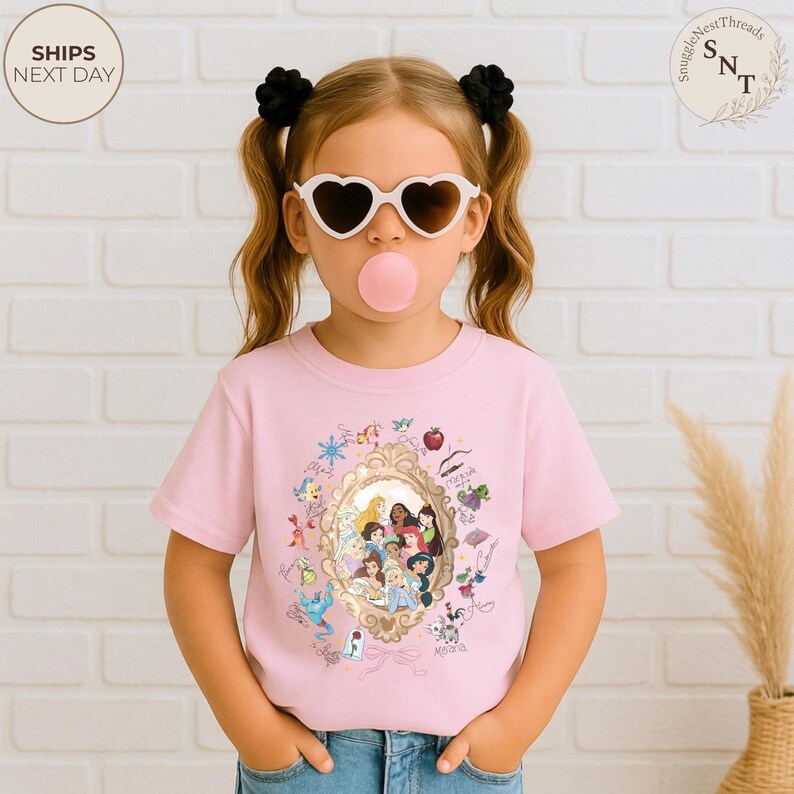 May include: A light pink t-shirt featuring a gold-framed illustration of Disney princesses. The shirt is worn by a child wearing heart-shaped sunglasses and a pink bubblegum bubble. The shirt has the text "SnuggleNestThreads" and "Ships Next Day".