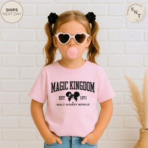 May include: A light pink t-shirt with the words "MAGIC KINGDOM" in black block letters. Below the text is "EST. 1971" and "WALT DISNEY WORLD". The shirt features a Mickey Mouse silhouette. The image shows a young person wearing heart-shaped sunglasses.