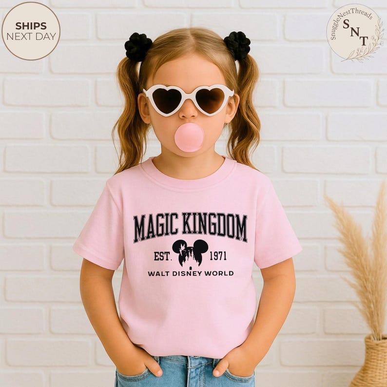 May include: A light pink t-shirt with the words "MAGIC KINGDOM" in black block letters. Below the text is "EST. 1971" and "WALT DISNEY WORLD". The shirt features a Mickey Mouse silhouette. The image shows a young person wearing heart-shaped sunglasses.