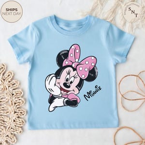 May include: A light blue t-shirt featuring a cartoon illustration of Minnie Mouse. Minnie Mouse is depicted with a pink bow with white polka dots, black ears, and a pink dress. The text "Minnie" is written in black cursive.
