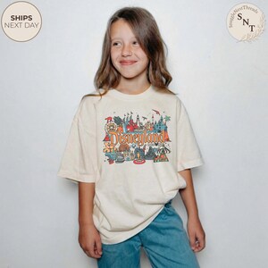 May include: A cream-colored t-shirt featuring a colorful Disneyland graphic. The graphic includes the Disneyland logo and various park icons. The shirt is worn by a person with long brown hair and blue jeans.