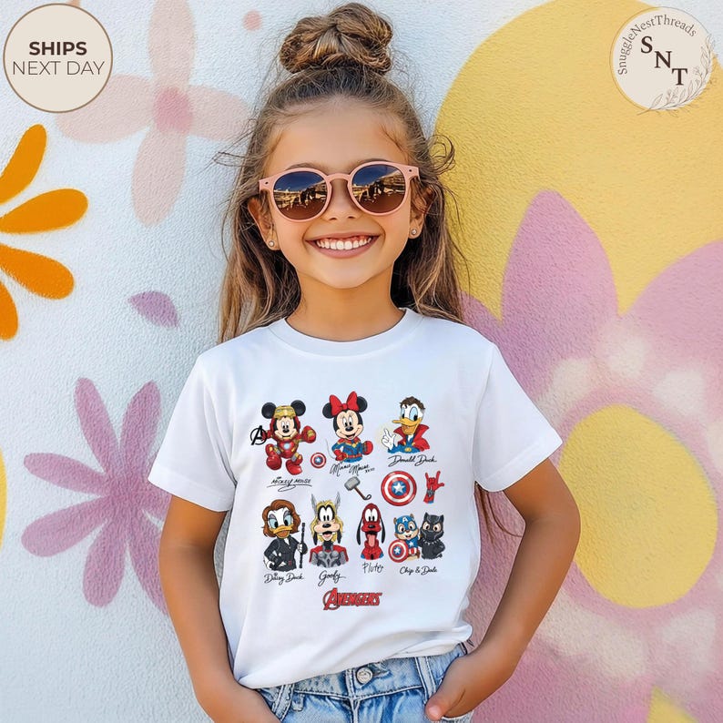 May include: A white t-shirt featuring colorful cartoon illustrations of Disney characters and Avengers symbols. The shirt includes Mickey Mouse, Minnie Mouse, Donald Duck, and other characters. The word "Avengers" is printed at the bottom.
