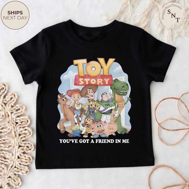 K&ouml;nnte beinhalten: Schwarzes T-Shirt mit einer farbenfrohen Grafik von Charakteren aus dem Film "Toy Story". Das Design umfasst Woody, Buzz Lightyear und andere Charaktere mit den Worten "TOY STORY" und "YOU'VE GOT A FRIEND IN ME."