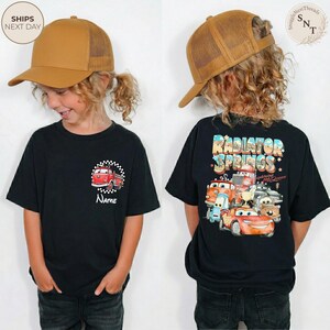 May include: Black t-shirt with a red and white cartoon bus graphic and the name "Name" on the front. The back of the shirt features a "Radiator Springs" design with various cartoon cars. The child is wearing a brown trucker hat.
