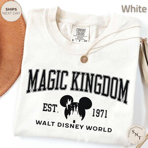 May include: White t-shirt with "MAGIC KINGDOM" in black block letters. Below the text is a black silhouette of Mickey Mouse ears with a castle, "EST. 1971", and "WALT DISNEY WORLD". A gold necklace with a pendant is on the shirt.