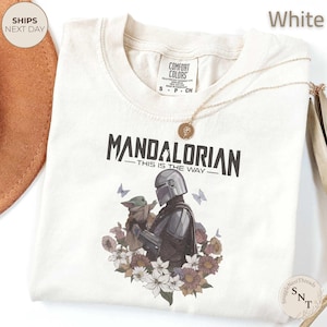 Puede incluir: Una camiseta blanca con un gr&aacute;fico del Mandaloriano sosteniendo a Grogu, rodeado de flores y mariposas. El texto dice "MANDALORIAN THIS IS THE WAY". La camiseta tiene una etiqueta Comfort Colors en el cuello y un collar con dije dorado.