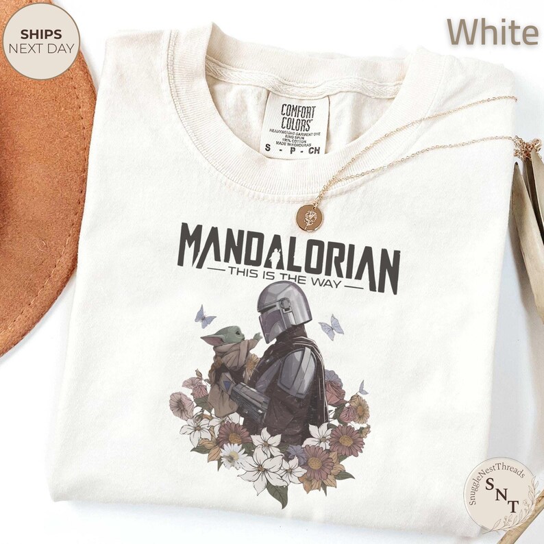 Puede incluir: Una camiseta blanca con un gr&aacute;fico del Mandaloriano sosteniendo a Grogu, rodeado de flores y mariposas. El texto dice "MANDALORIAN THIS IS THE WAY". La camiseta tiene una etiqueta Comfort Colors en el cuello y un collar con dije dorado.