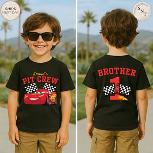 May include: A black t-shirt with a red race car graphic and the text "David's PIT CREW" on the front. The back of the shirt says "BROTHER 1" with a race car and checkered flags. The boy is wearing sunglasses and khaki shorts.