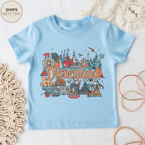 May include: A light blue t-shirt featuring a colorful graphic of Disneyland Resort. The design includes various park icons and the words "Disneyland Resort" in orange and red. The shirt has short sleeves and a crew neck.