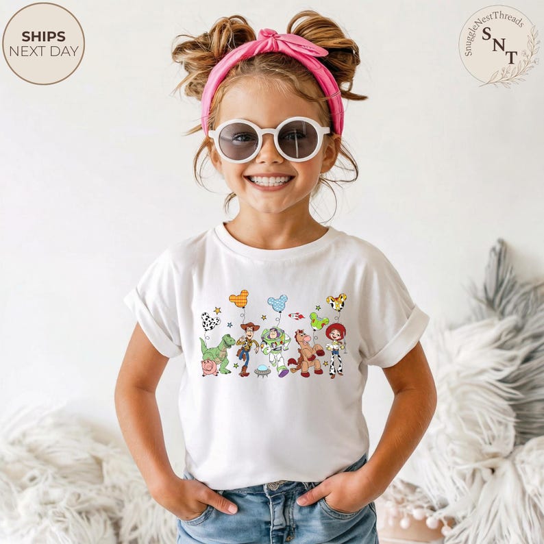 May include: A white t-shirt featuring a colorful graphic of characters from the Toy Story movie. The design includes images of a dinosaur, Woody, Buzz Lightyear, and other characters. The shirt is worn by a young person wearing sunglasses and a pink headband.