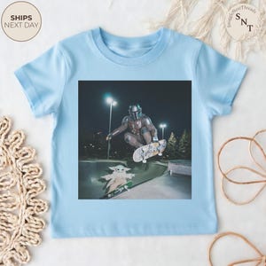 May include: Light blue t-shirt featuring a graphic of a character from the Mandalorian series skateboarding in a skate park. The image includes the character and Grogu on skateboards. The shirt has a crew neck and short sleeves.