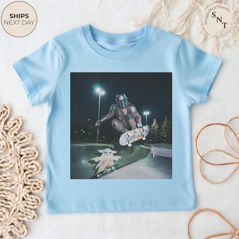 May include: Light blue t-shirt featuring a graphic of a character from the Mandalorian series skateboarding in a skate park. The image includes the character and Grogu on skateboards. The shirt has a crew neck and short sleeves.